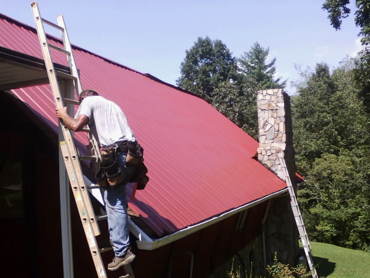 Roofer on a ladder beside a red metal roof and stone chimney — Soffit & Fascia Repair by Roofing Brothers in Bottineau